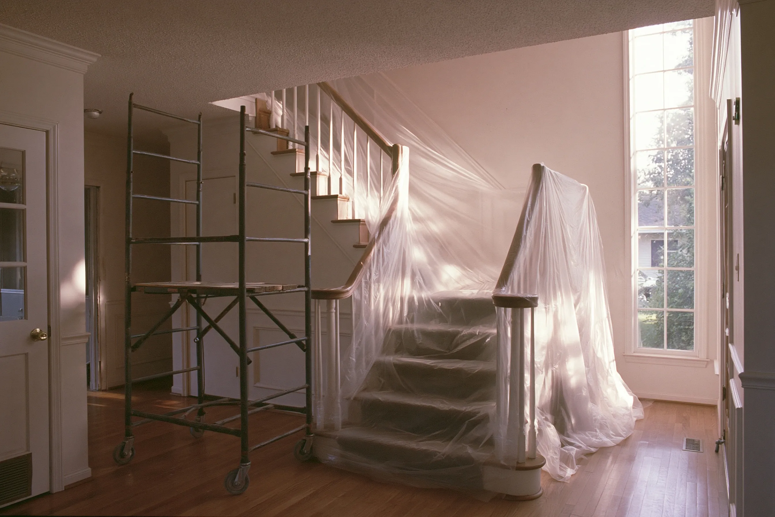 High textured ceiling above a staircase, a rolling scaffold at the foot of the stairs and the steps and rail wrapped in plastic
